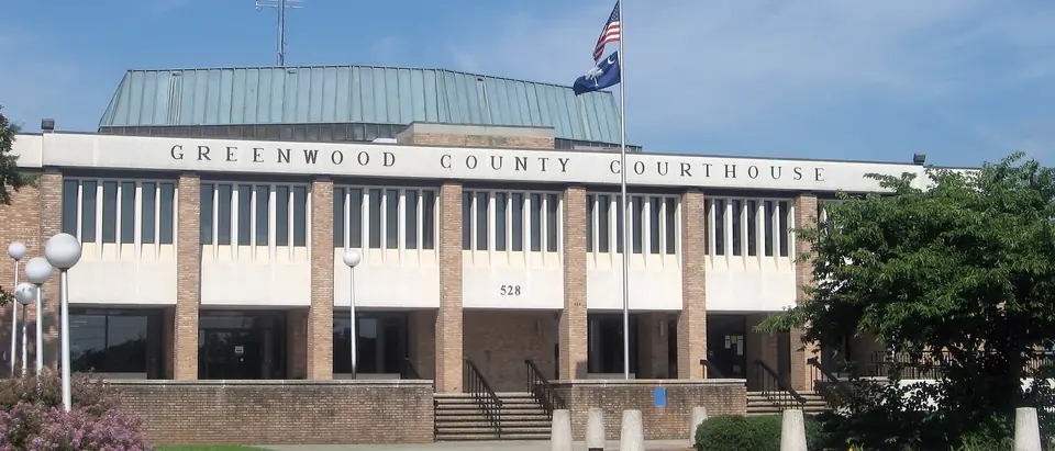 The Greenwood County Courthouse with a flag and crape myrtle under a blue sky in Greenwood, SC