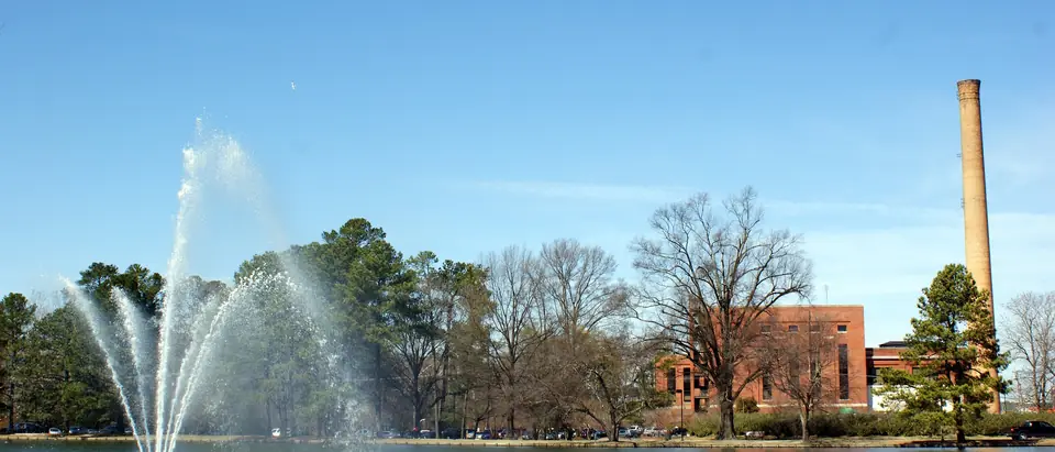 A fountain in City Lake with the old mill chimney and brick buildings behind, in Rocky Mount, NC