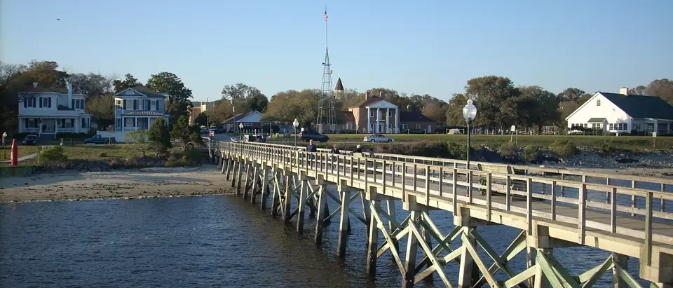 A wooden fishing pier leading back to the waterfront houses of Southport, NC, under a clear blue sky