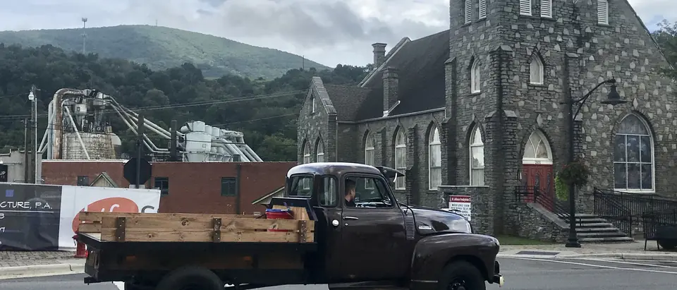A vintage pickup truck passing a stone church downtown with Mount Jefferson behind, in West Jefferson, NC