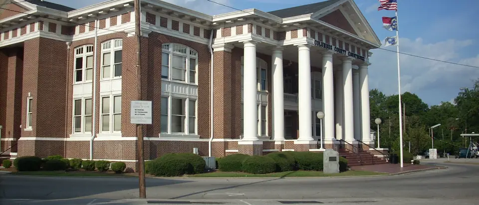 The white-columned Columbus County Courthouse with flags under a blue sky in Whiteville, NC